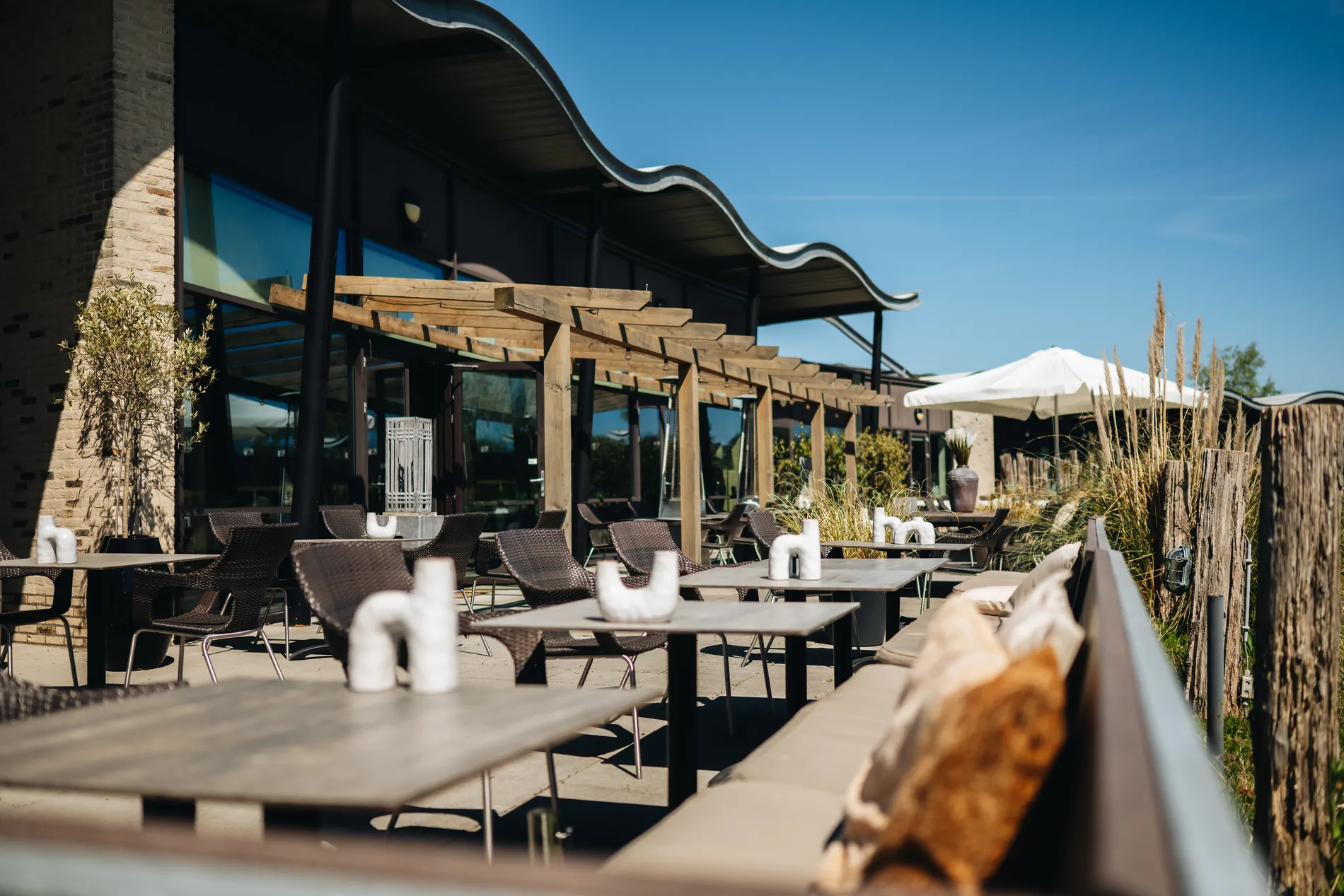 Het lege, zonnige terras van het restaurant met houten pergola's, moderne tafels en stoelen, en een opvallend golvend dakontwerp.